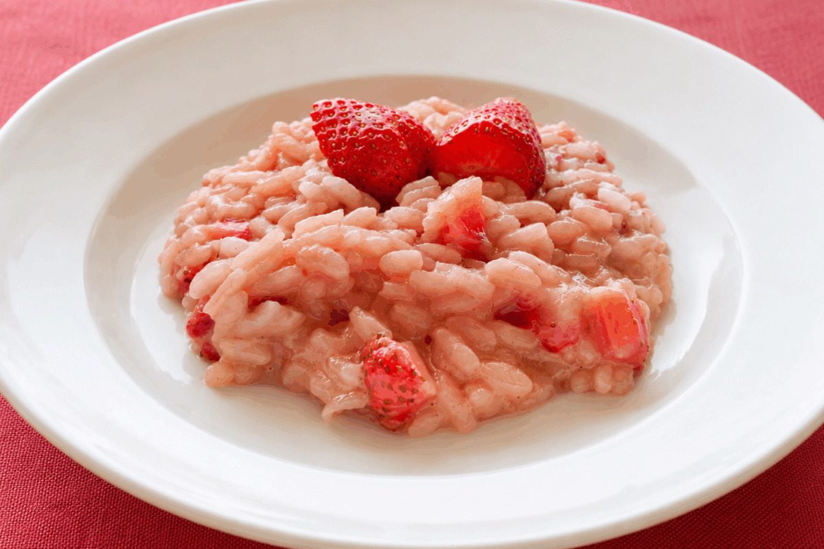Piatto bianco con risotto alle fragole cremoso in versione senza lattosio e vegana, servito su una tovaglia di lino rosso con fragole fresche in superficie
