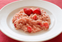 Piatto bianco con risotto alle fragole cremoso in versione senza lattosio e vegana, servito su una tovaglia di lino rosso con fragole fresche in superficie