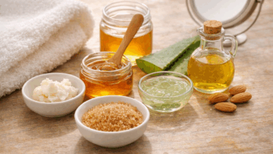 Ingredienti naturali per la cura delle labbra su tavolo in legno chiaro: miele, burro di karité, aloe vera, zucchero di canna e olio di mandorle con asciugamano bianco in luce naturale