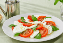 Insalata caprese servita su un grande piatto bianco con pomodoro, mozzarella e basilico, appoggiata su una tovaglia verde chiaro, con oliera in lattina, bicchiere d’acqua con ghiaccio e ciotola di taralli a fianco