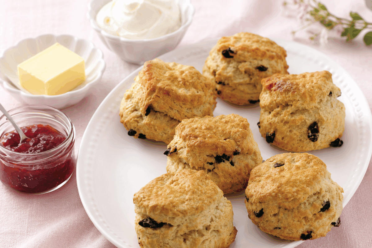 Scones dorati serviti su un piatto bianco con vasetto di marmellata di fragole, panetto di burro e ciotola di panna montata su tovaglia di lino rosa pallido