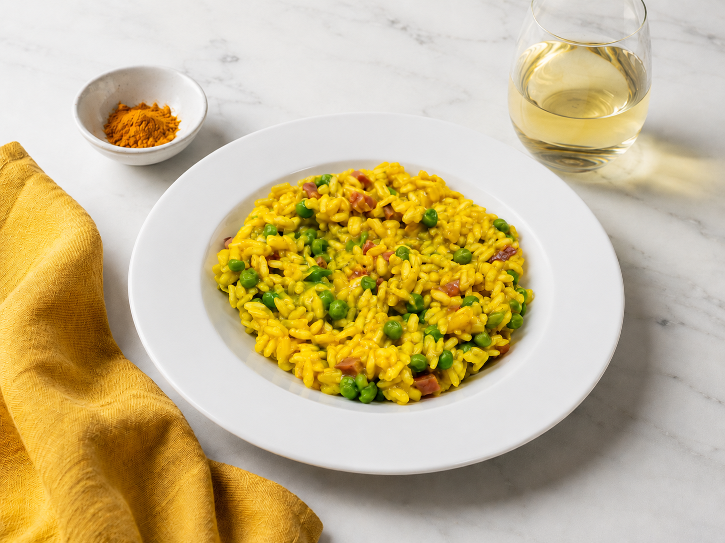 Piatto bianco di porcellana con risotto alla curcuma, carote e piselli su tavolo di marmo bianco, con tovagliolo di lino giallo senape, ciotolina di curcuma e bicchiere di vino bianco