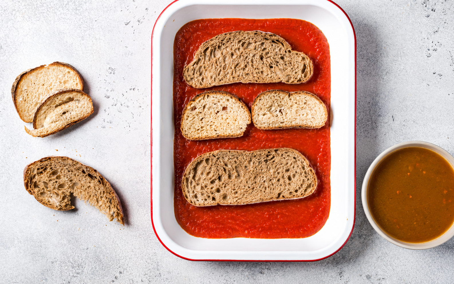 Pirofila con fette di pane raffermo disposte su passata di pomodoro, accanto pane casereccio e una ciotola di minestrone passato liquido, vista dall’alto.