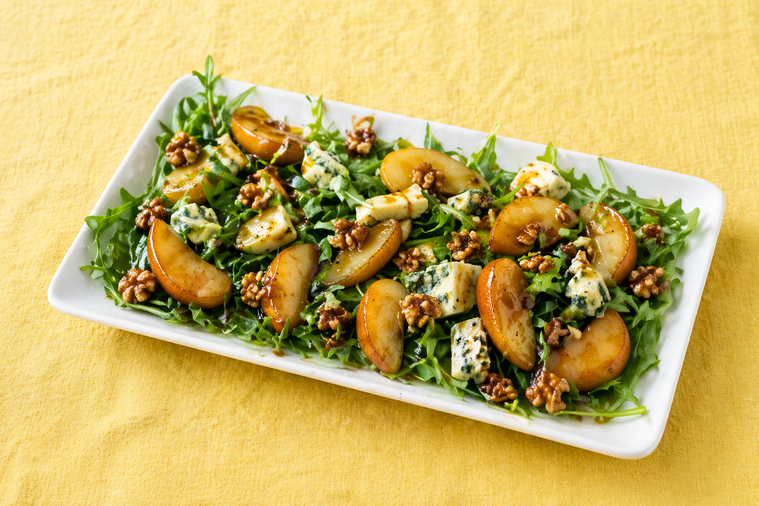 Insalata di valeriana con pere caramellate, noci e gorgonzola servita in piatto rettangolare bianco su tovaglia di lino giallo