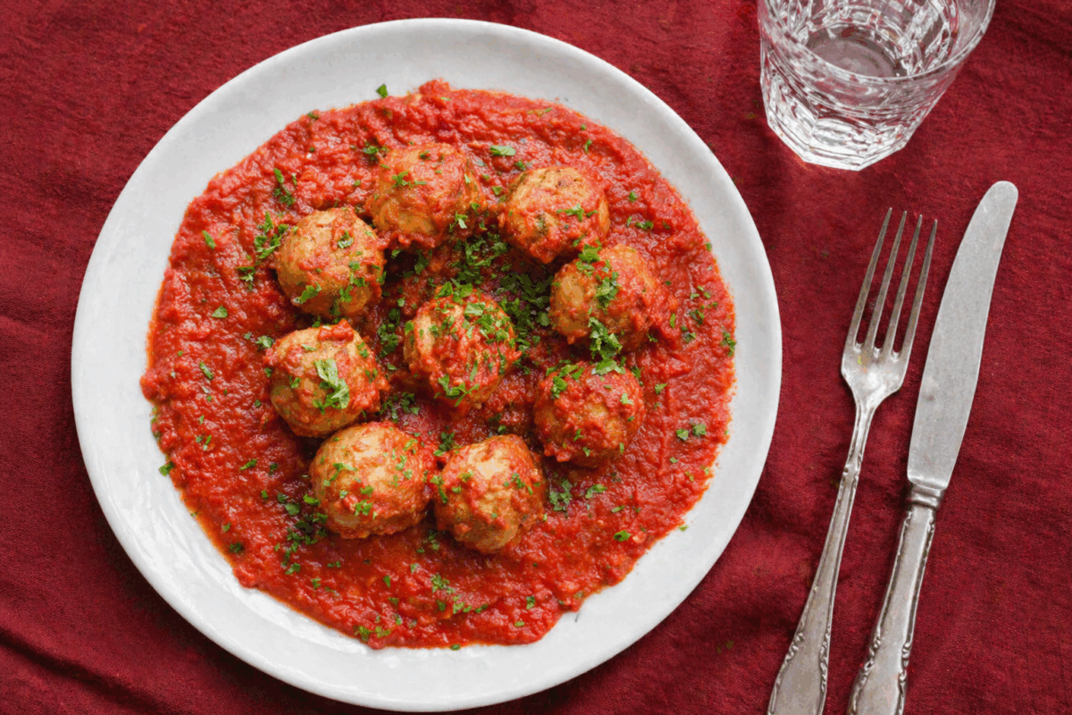 Polpette al sugo servite in piatto bianco su tovaglia di lino bordeaux con posate e bicchiere d’acqua, vista dall’alto