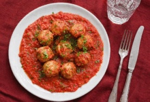 Polpette al sugo servite in piatto bianco su tovaglia di lino bordeaux con posate e bicchiere d’acqua, vista dall’alto