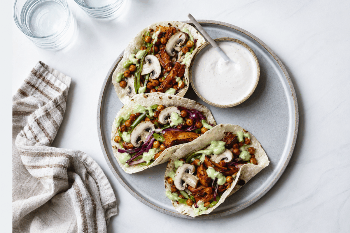 tre tacos vegetariani con funghi champignon, ceci croccanti e salsa verde su un piatto grigio, accompagnati da una ciotola di salsa bianca, due bicchieri d’acqua e un canovaccio in lino a righe marroni.