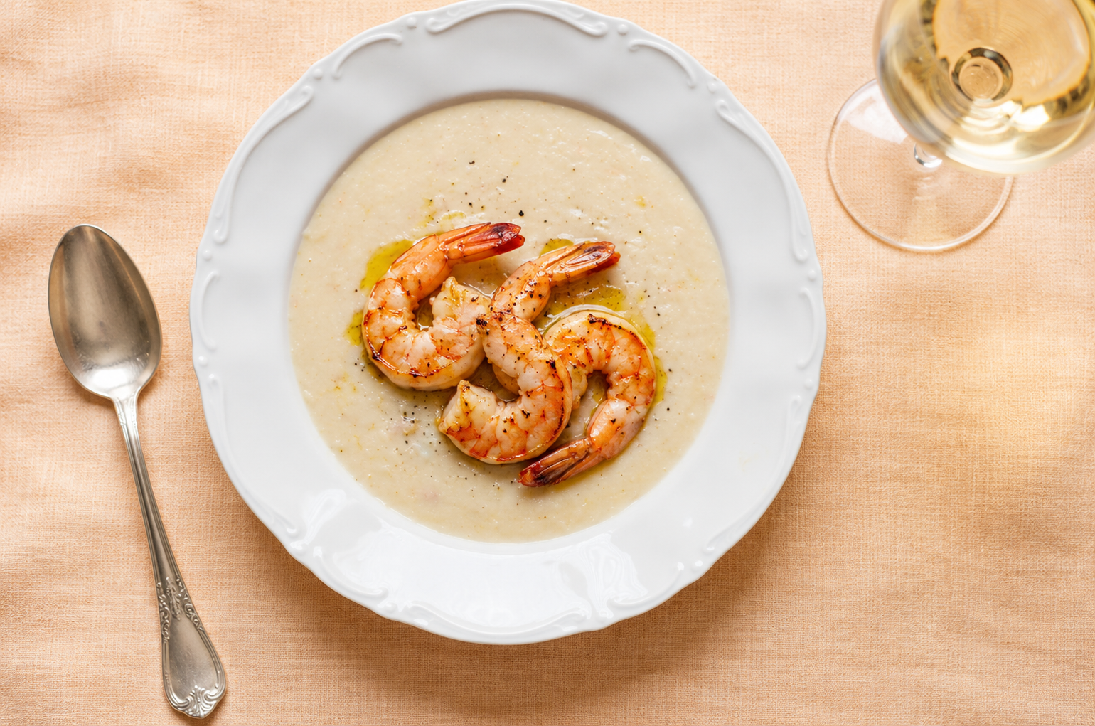 Vellutata di ceci servita in un piatto bianco con scampi grigliati, su tovaglia di lino arancione pallido, con cucchiaio d’argento e calice di vino bianco