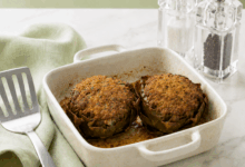 Carciofi ripieni al forno in una casseruola di ceramica leggermente sporca dalla cottura, con sugo e grasso sul fondo, su tavolo di marmo bianco con tovagliolo verde pallido, paletta in acciaio e macinini trasparenti per sale e pepe