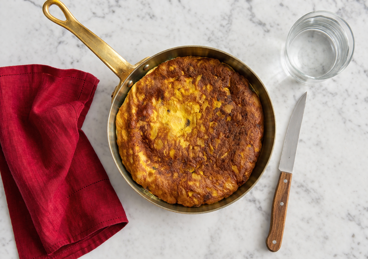 Frittata dorata in padella vista dall’alto su piano in marmo bianco con tovagliolo rosso, coltello e bicchiere d’acqua