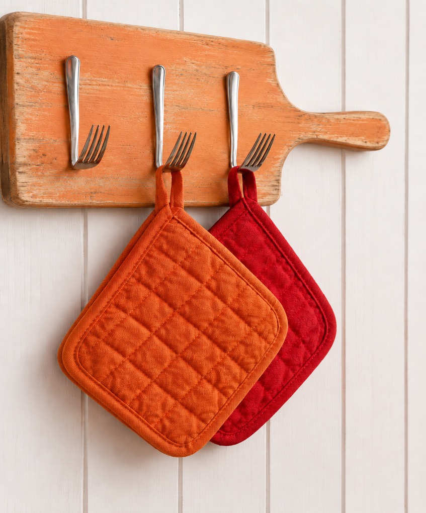 Tagliere di legno arancione trasformato in appendipresine fai da te, con forchette riciclate usate come ganci e presine arancioni e rosse appese su parete bianca a doghe