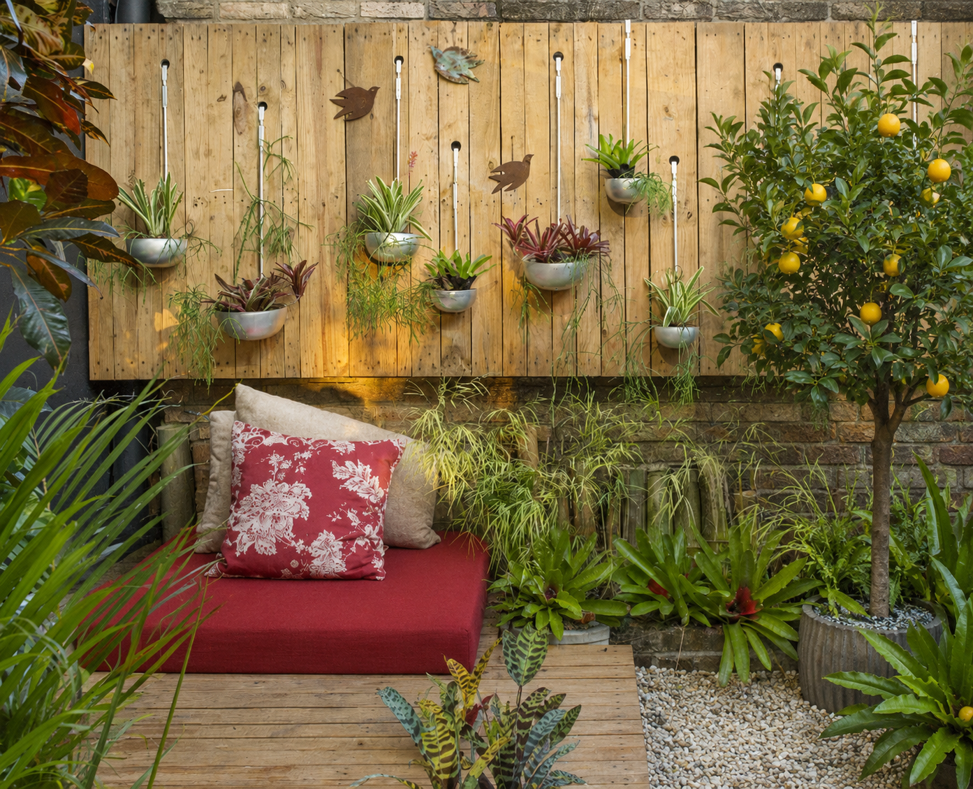 Giardino eclettico e bohémien con pedana in legno, grande cuscino rosso, parete verde con vasi sospesi, ghiaia e albero di agrumi