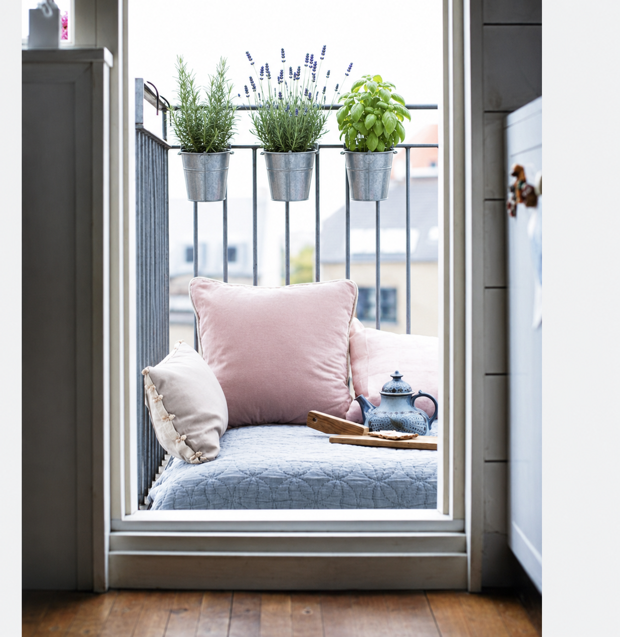 Mini-balcone arredato come angolo relax per la bella stagione, con cuscino rosa pallido, coperta chiara, vassoio da tè e tre vasetti di aromatiche appesi alla ringhiera, ideale per vivere all’aperto anche in poco spazio