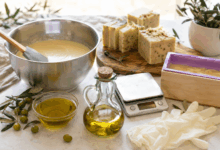 Preparazione del sapone naturale all’olio d’oliva vista da vicino, con ciotola in acciaio, olio d’oliva, stampo, panetti di sapone, bilancia e guanti protettivi su un piano luminoso