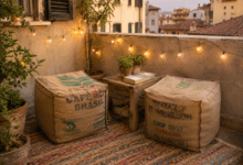 Due pouf fai-da-te realizzati con vecchi sacchi di juta da caffè, sistemati su un terrazzino con tappeto colorato, tavolino in legno, libro, piante grasse e luminarie davanti a case di una città italiana