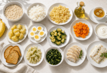 Tavola imbandita con piatti bianchi contenenti i cibi più adatti a chi soffre di ernia iatale, tra cui avena cotta, riso in bianco, pasta semplice, patate lesse, pane tostato, uova sode, yogurt magro, banana, pesce bianco, pollo, zucchine, carote, finocchi e bietole cotte
