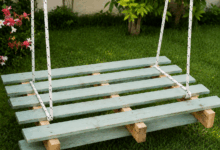 Altalena rustica realizzata con pallet di legno verniciato verde chiaro, appesa con corde bianche in un giardino curato con prato e piante fiorite sullo sfondo