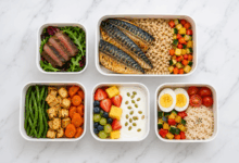 cinque bento box aperti su piano di marmo bianco, con bistecca e insalata, sgombro con orzo e verdure, tofu con fagiolini e carote, frutta con yogurt e semi di zucca, uova sode con riso e ortaggi.