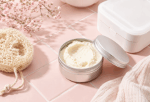 Vasetto di metallo con burro di karité su piastrelline rosa pallido, con scatolina bianca e spugna corpo di loofah in stile flat-lay elegante