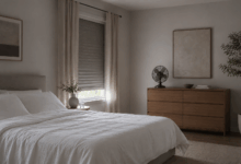Camera da letto minimal in penombra con lenzuola bianche di cotone leggero, tapparella quasi abbassata, pochi mobili di design e ventilatore su cassettiera