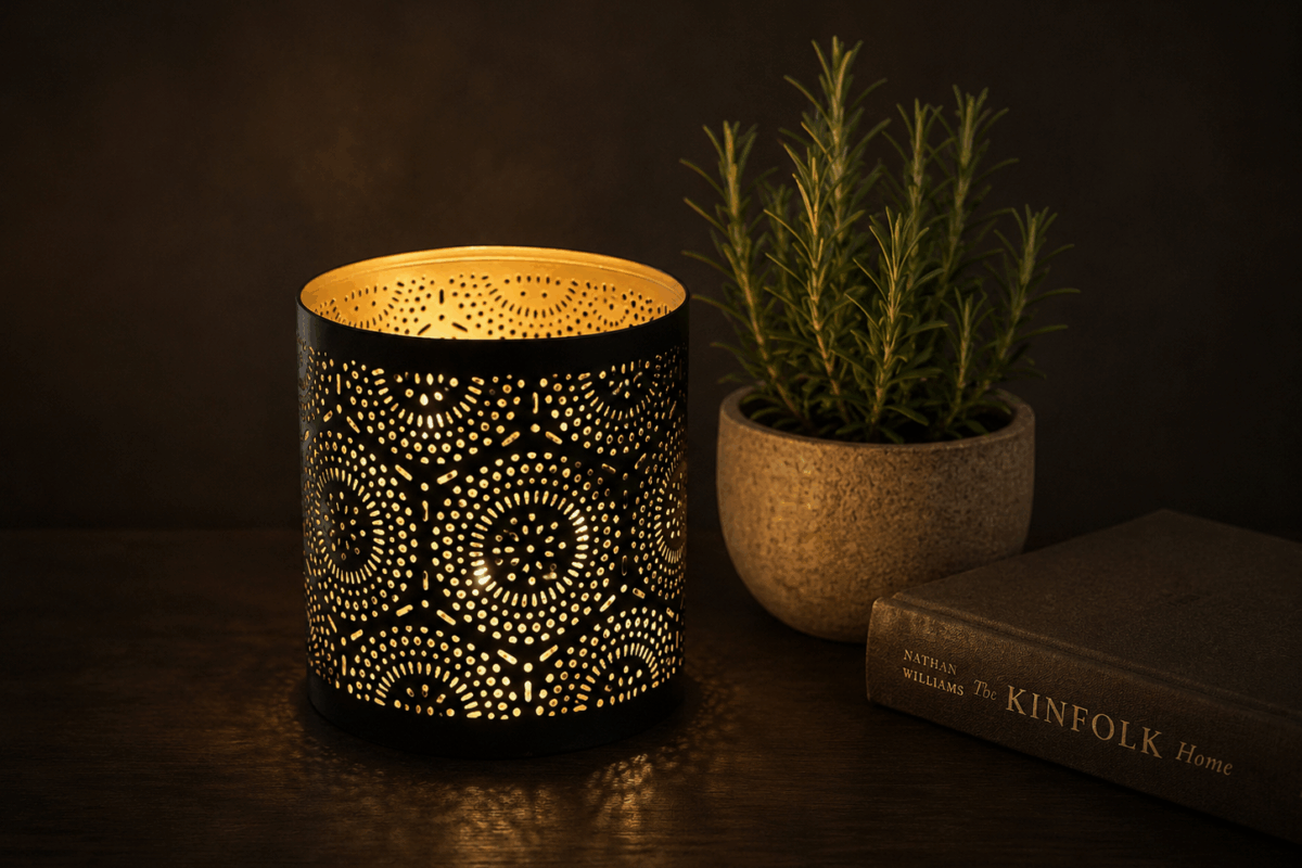 Candela decorativa traforata accesa su tavolo scuro, accanto a un vasetto di rosmarino e a un libro, con sfondo uniforme e soffuso
