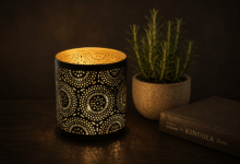 Candela decorativa traforata accesa su tavolo scuro, accanto a un vasetto di rosmarino e a un libro, con sfondo uniforme e soffuso