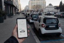 Mano che tiene uno smartphone con app di car sharing aperta, davanti ad auto compatte parcheggiate lungo una strada urbana italiana