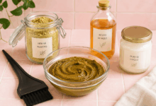 Ciotola di vetro con pasta di henné per capelli su piastrelle rosa chiaro, con pennello piatto da tinta, barattolo di henné neutro, bottiglietta di aceto di mele e vasetto di olio di cocco con etichette minimal