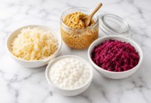 Tre cibi fermentati comuni su piano in marmo bianco: kefir in ciotola, miso in barattolo trasparente e crauti bianchi e rossi in ciotole.
