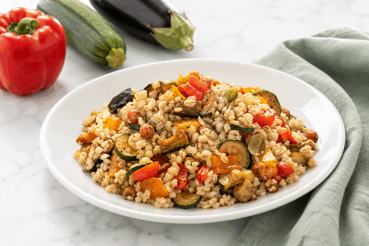 Piatto di porcellana bianca con insalata di riso e verdure estive su superficie di marmo bianco, con tovagliolo in lino verde salvia e accanto un peperone rosso, una zucchina e una melanzana