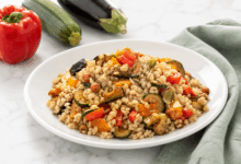 Piatto di porcellana bianca con insalata di riso e verdure estive su superficie di marmo bianco, con tovagliolo in lino verde salvia e accanto un peperone rosso, una zucchina e una melanzana