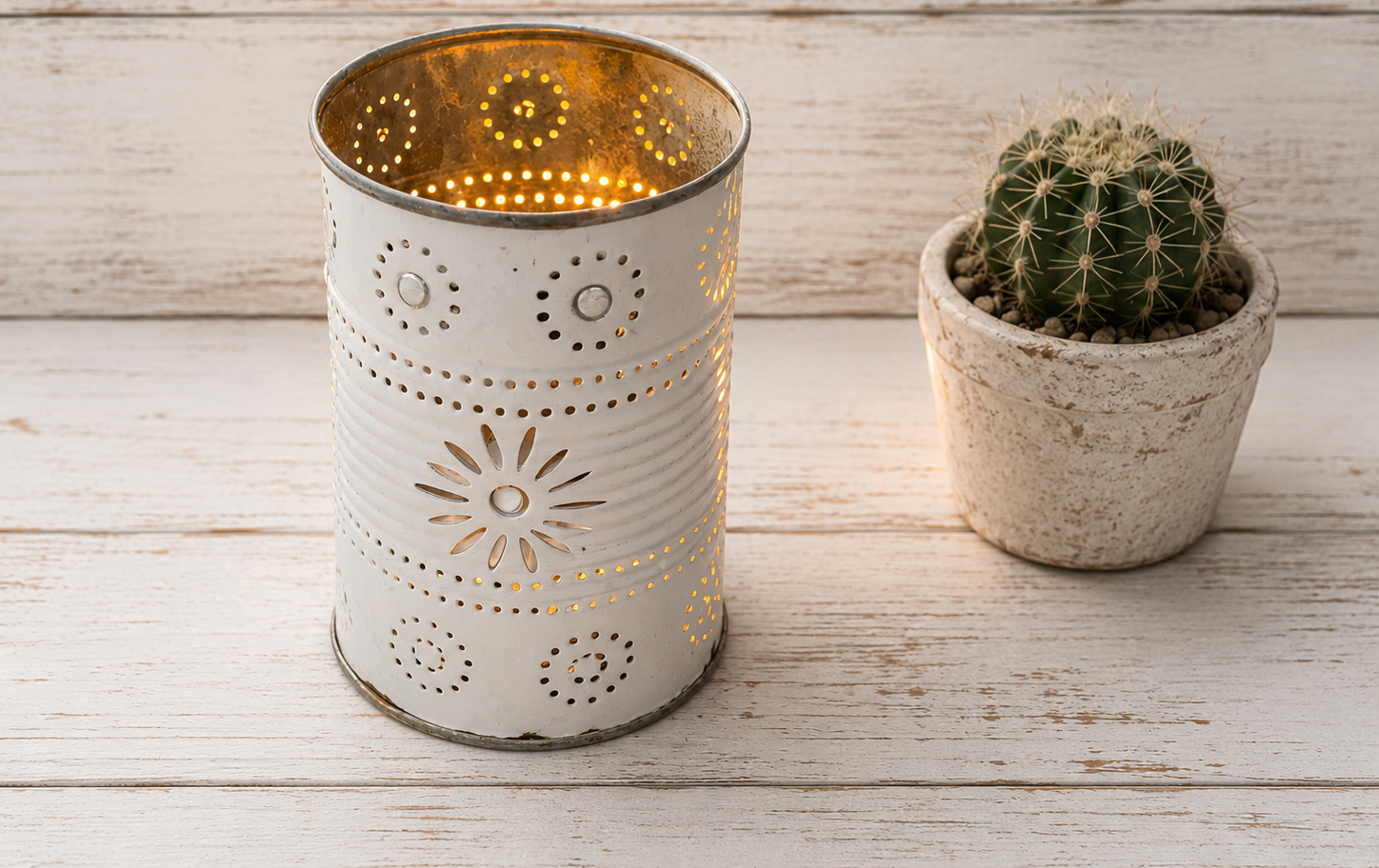 Portacandela in latta bianca traforata acceso su sfondo di legno bianco decapato, accanto a un piccolo vaso di cactus