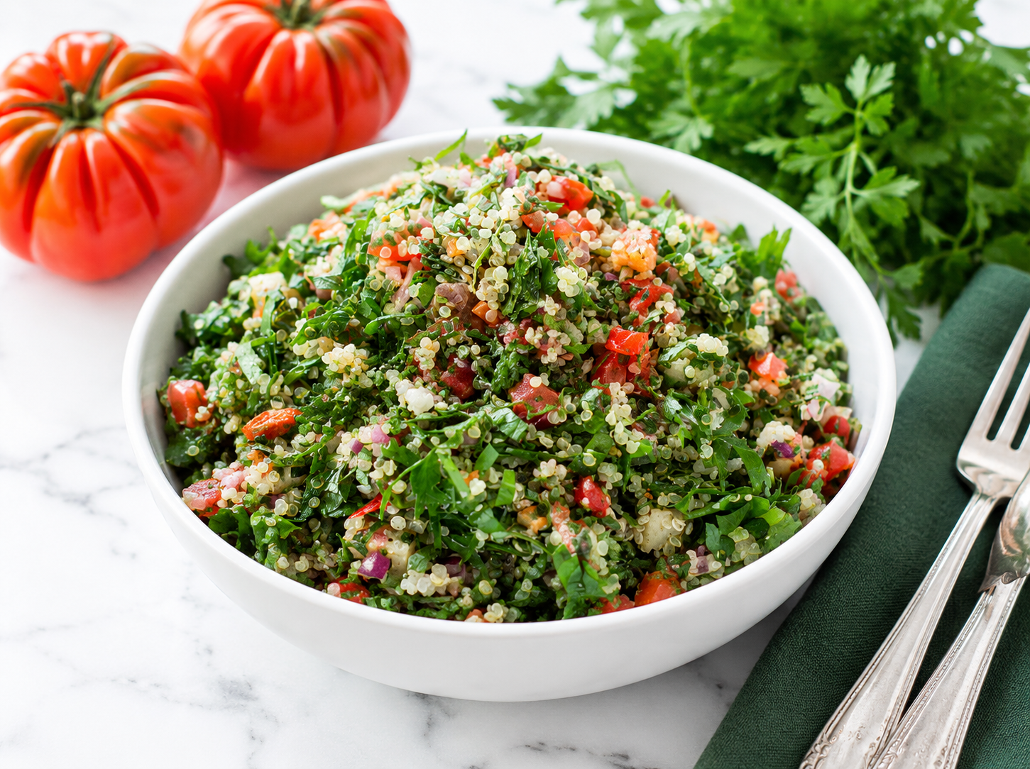 Taboulé servito in un piatto bianco su tavolo di marmo bianco, con prezzemolo, pomodori e cipollati a rondelle, accanto a un tovagliolo di lino verde scuro e posate d’argento