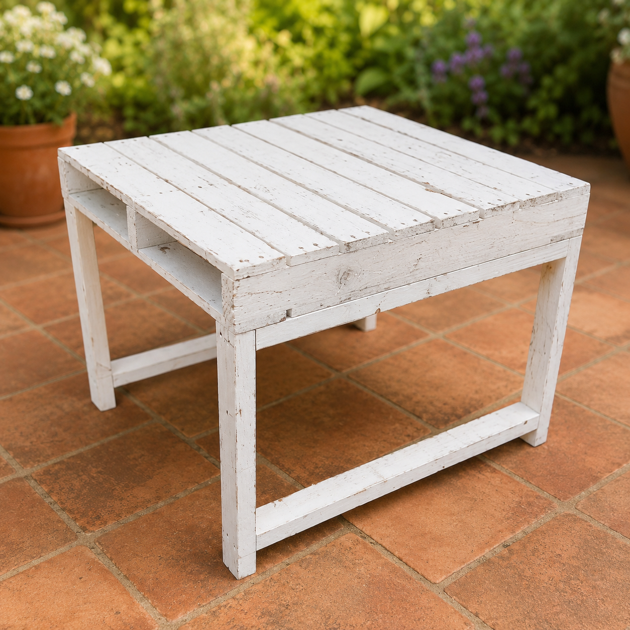Tavolino in pallet dipinto di bianco con finitura rustica, su pavimento in cotto in un patio con giardino sullo sfondo