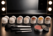 Barattoli di ingredienti minerali per trucco fai da te, tra cui amido di riso, caolino bianco, argilla rosa, mica e ossidi colorati, allineati su un piano nero davanti a uno specchio da makeup con luci, con pennelli e blush minerale già pronto