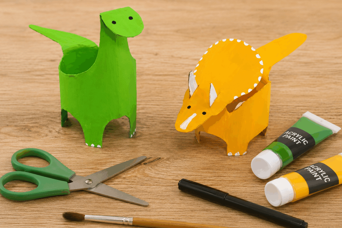 Due dinosauri realizzati con rotoli di carta igienica riciclati e dipinti di verde e giallo, appoggiati su un piano di legno con forbici, pennello, pennarello nero e colori acrilici