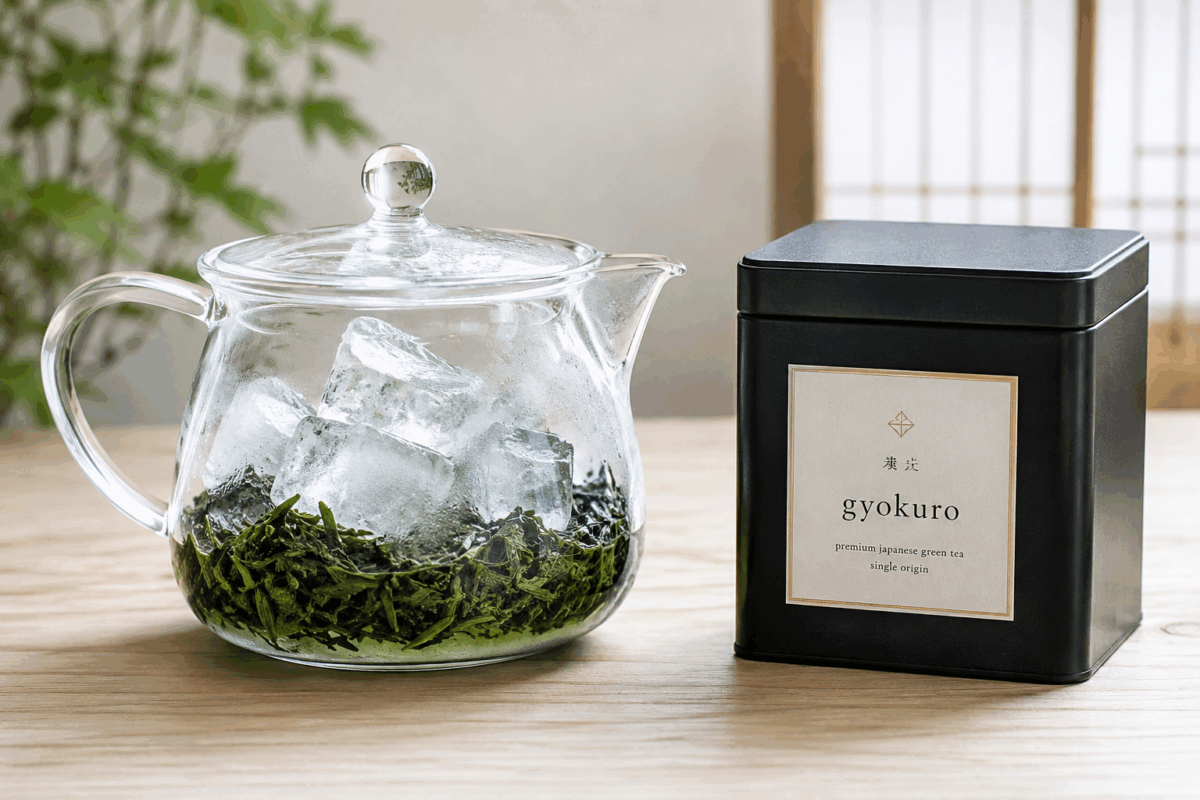 Teiera di vetro con foglie di tè verde giapponese coperte da cubetti di ghiaccio per la preparazione kōridashi, accanto a una scatola nera di gyokuro su un tavolo di legno chiaro